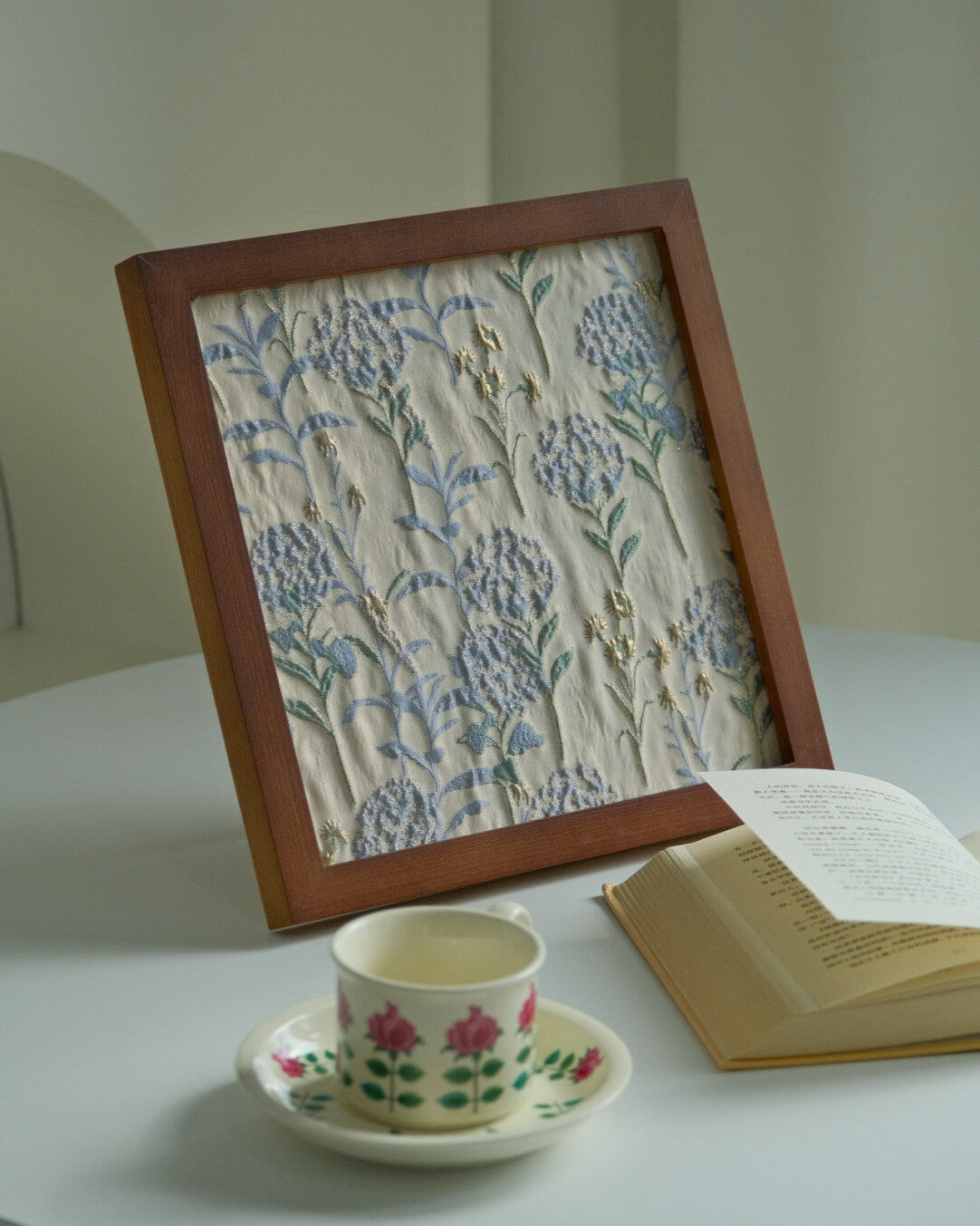 Hydrangea floral framed fabric art standing on a table with a teacup and open book