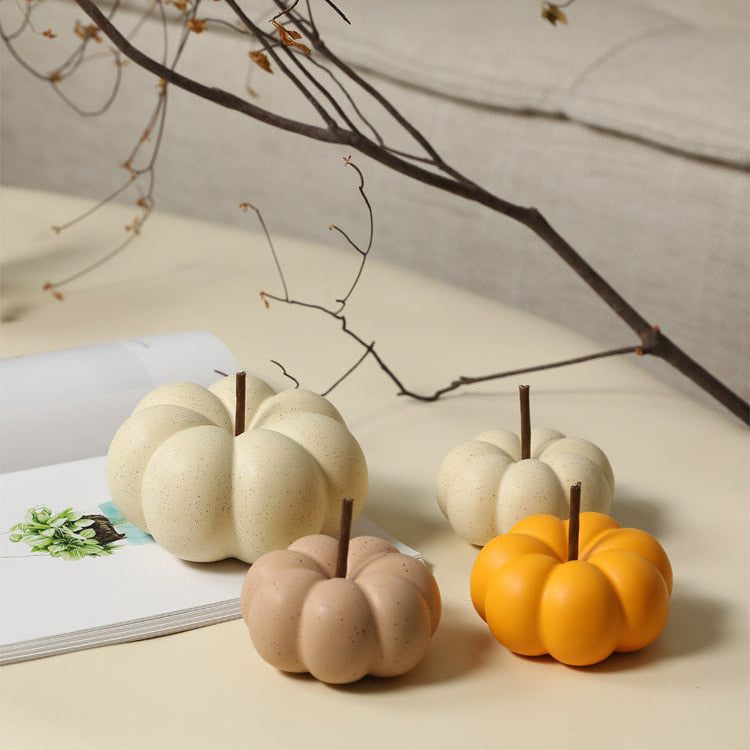 Four small decorative pumpkin-shaped objects in cream, tan, and orange, arranged on a light beige tabletop with a book and bare branches in the background.