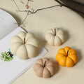 Three decorative faux pumpkin ornaments in cream, peach, and orange-yellow, arranged on a beige tabletop next to an open book with a branch in the background.
