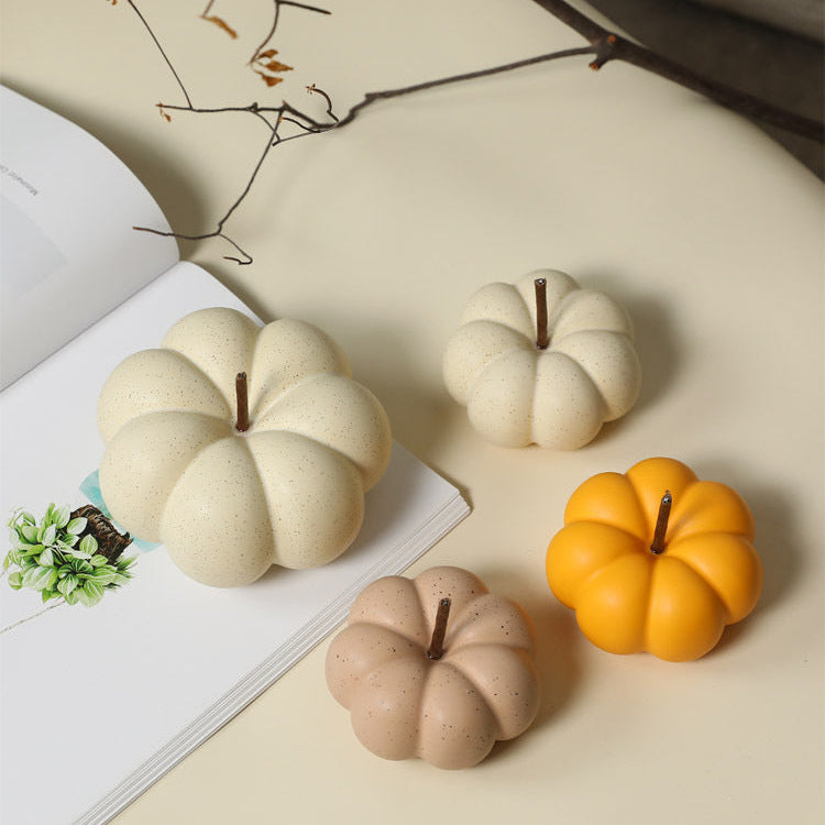 Three decorative faux pumpkin ornaments in cream, peach, and orange-yellow, arranged on a beige tabletop next to an open book with a branch in the background.