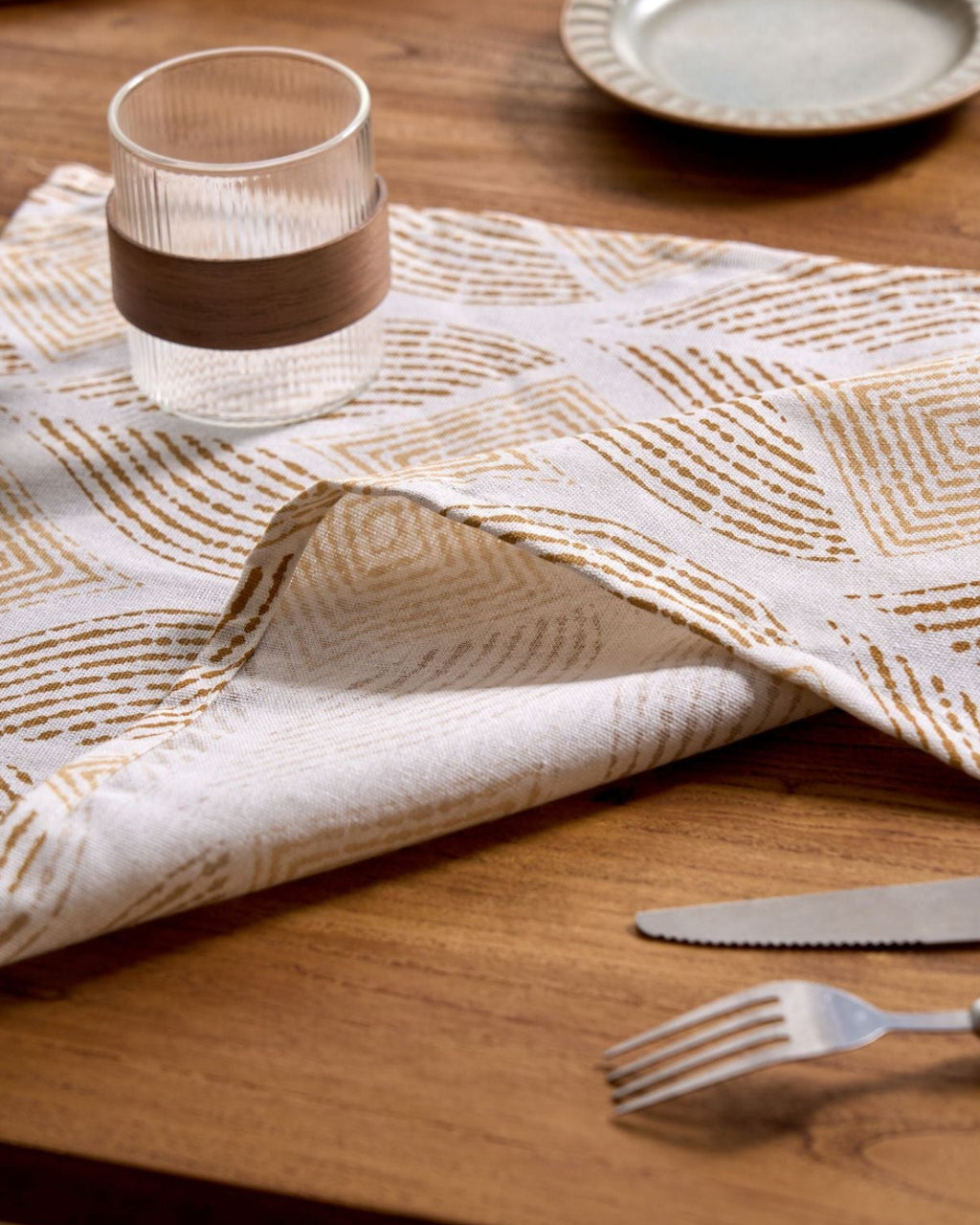 Table setting with a patterned cloth, glass, and cutlery on a wooden table.