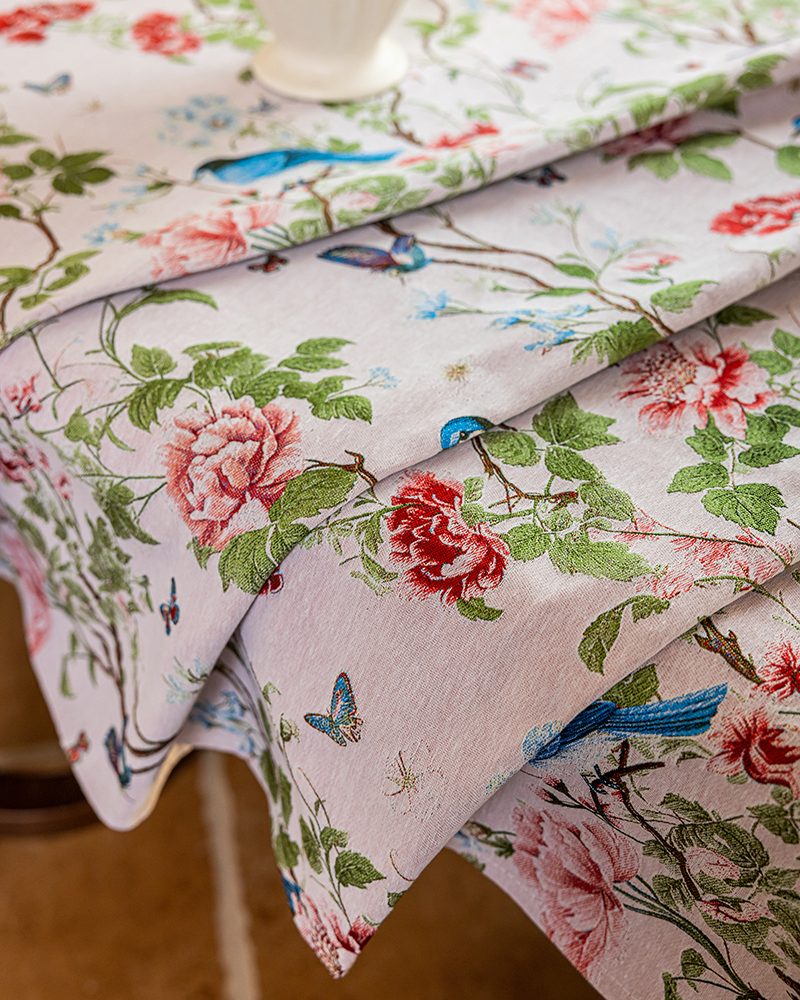 A close-up of a draped tablecloth with a classic toile-style print featuring pink and red flowers, green leaves, blue birds, and butterflies on a light background.