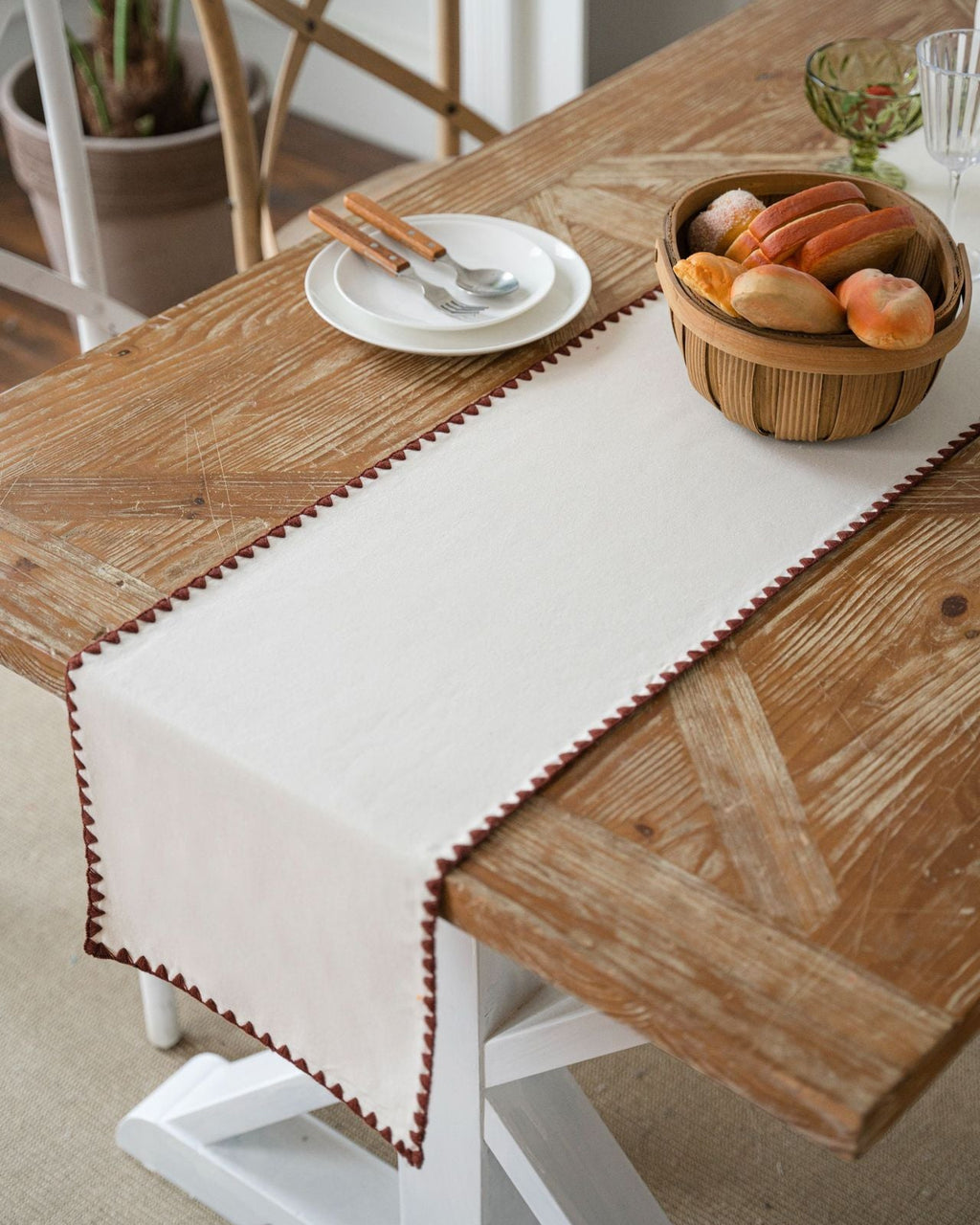 Wooden dining table with a white table runner, plates, cutlery, and a basket of fruits.