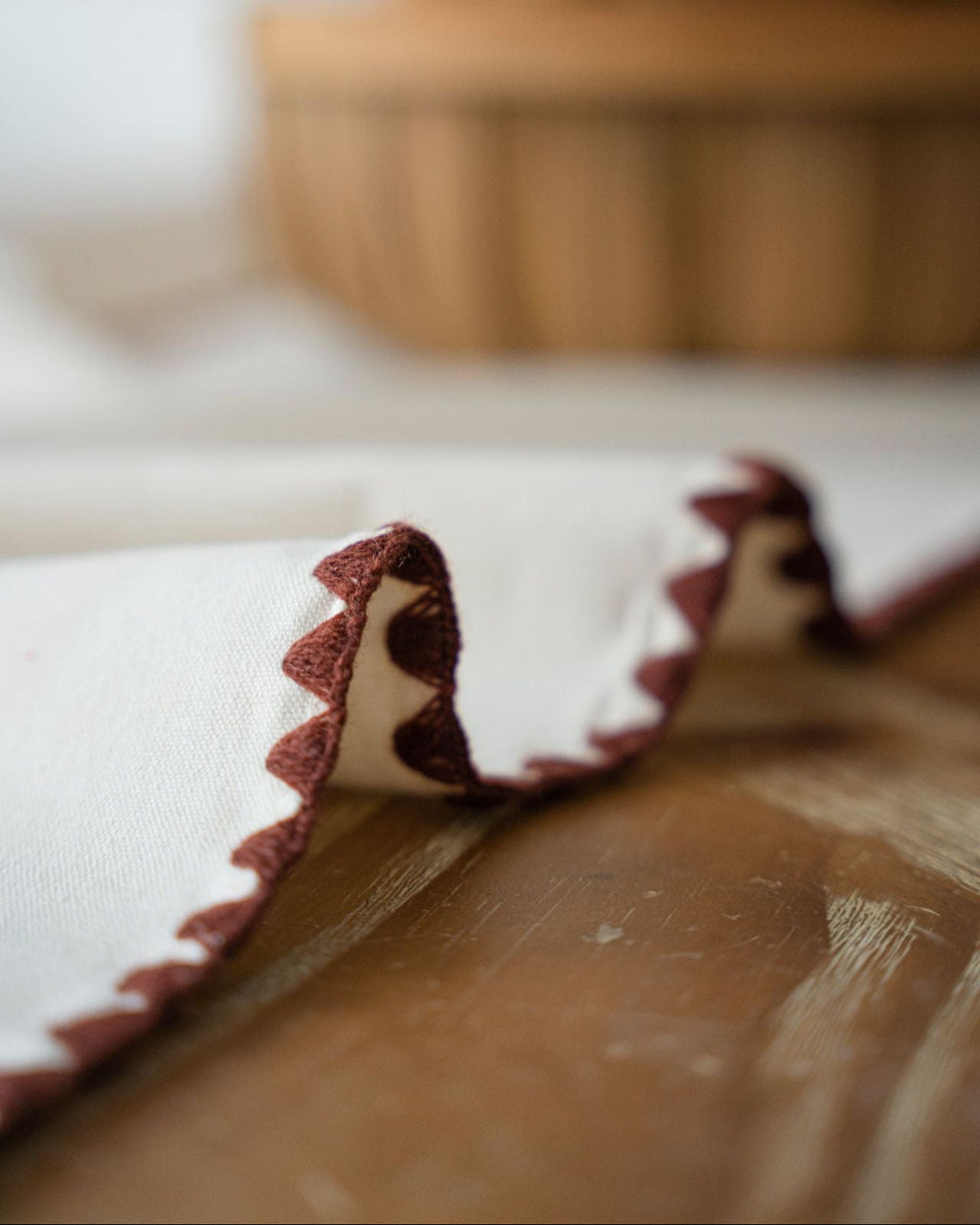 Close-up of a white napkin with brown trim on a wooden surface, blurred basket in the background.