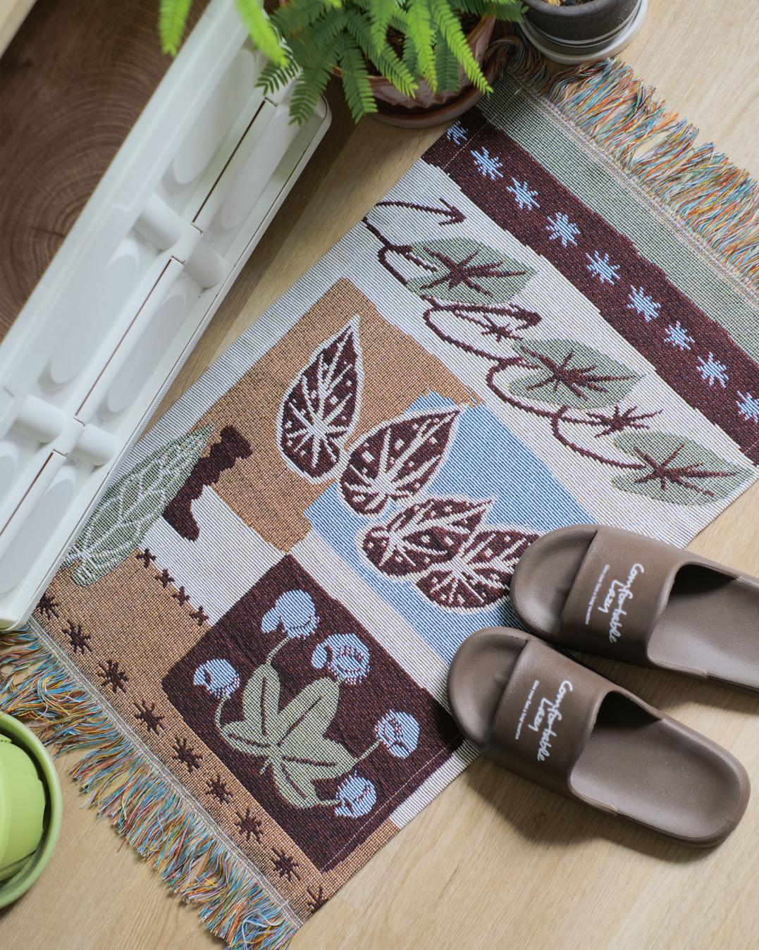 Woven fringe tapestry laid on the floor beside slippers and a plant stand